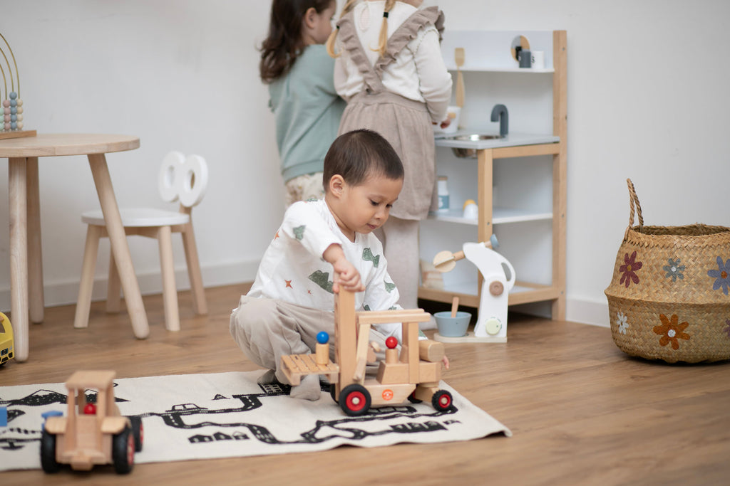 Kleiner Junge spielt in Kinderecke auf Spielzeugteppich mit Holz Gabelstapler und zwei Mädchen im Hintergrund stehen an einer Kinderküche aus Holz in weiß
