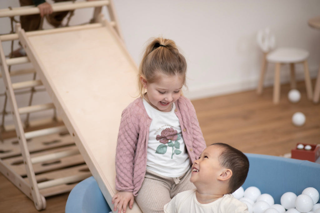 zwei Kinder lachen sich gegenseitig an in einem blauen Bällebad mit Kletterdreieck und Rutsche im Hintergrund