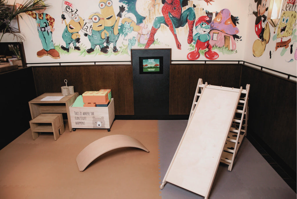 Kinderecke im Café Extrablatt mit Kletterdreieck, Rutschbrett, Balancierbrett aus Holz, Kindertisch und Hocker, und Aufbewahrungsbox mit großen Bausteinen auf Spielmatten