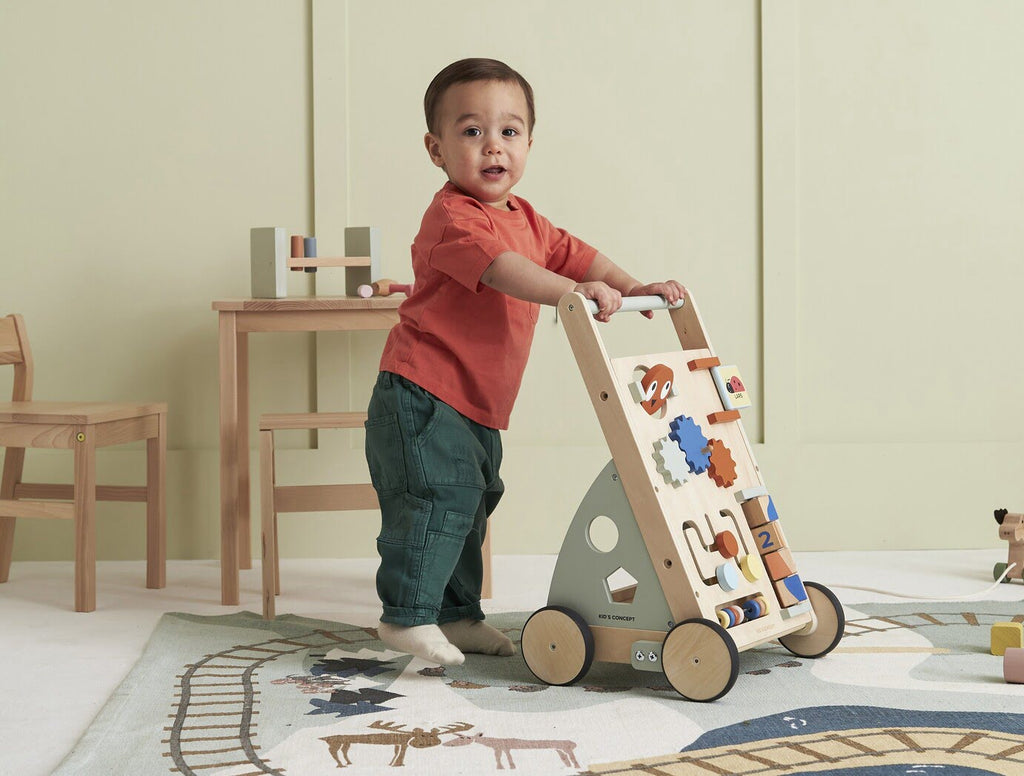 Kind mit Lauflernwagen aus Holz mit Activitybrett auf Spielteppich im Kinderzimmer