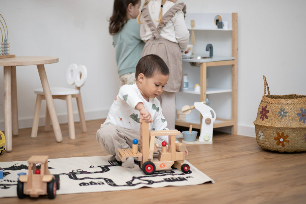 Junge spielt auf Spielteppich mit Gabelstapler aus Holz und zwei Mädchen spielen in Kinderküche im Hintergrund