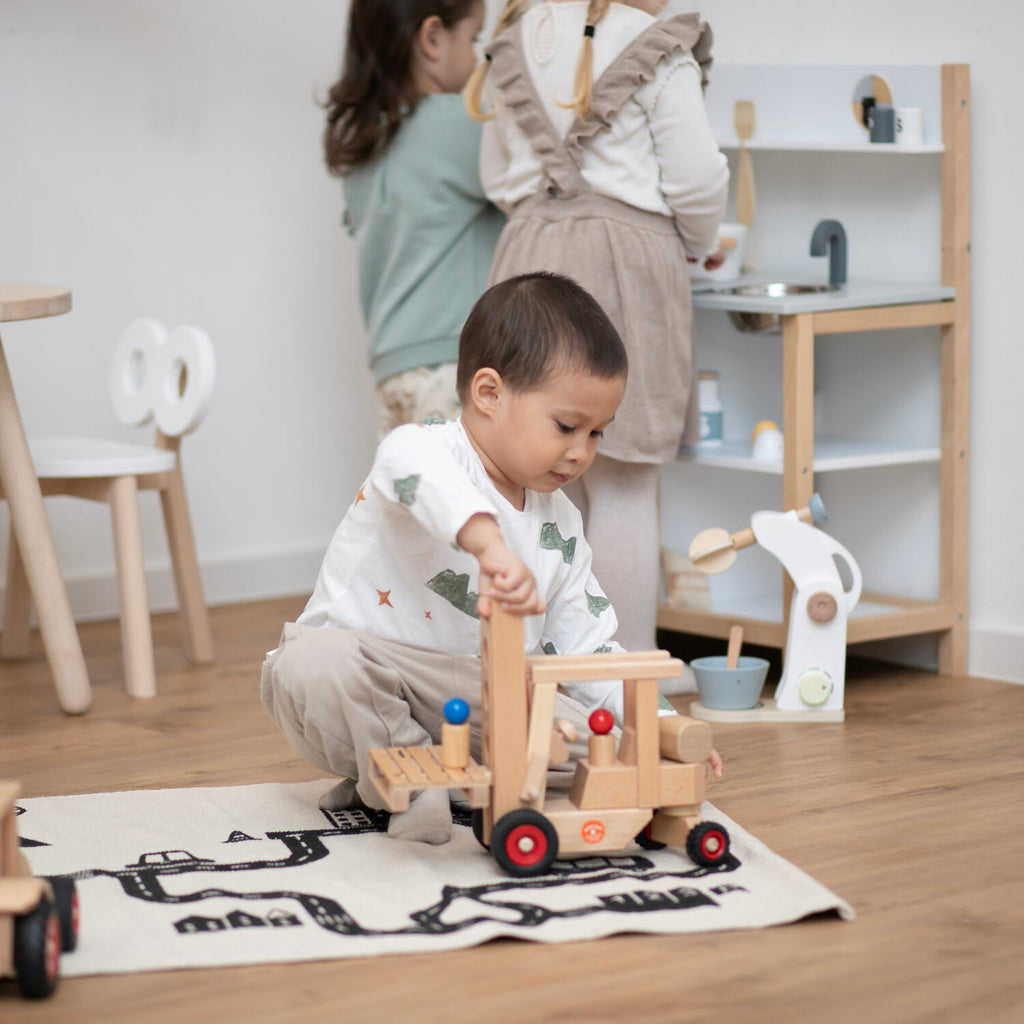 Kleiner Junge spielt in Kinderecke auf Spielzeugteppich mit Holz Gabelstapler und zwei Mädchen im Hintergrund stehen an einer Kinderküche aus Holz in weiß