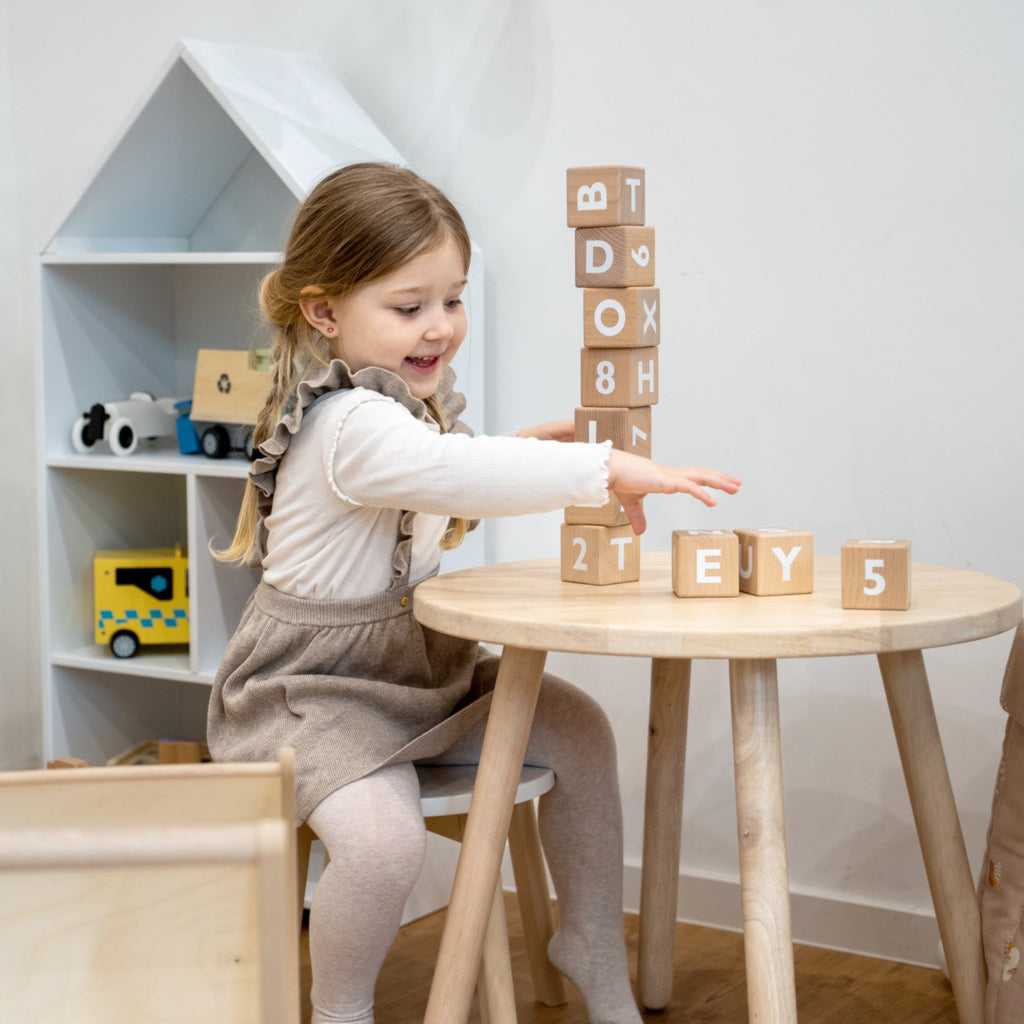 Mädchen sitzt am runden Kindertisch und stapelt Holz Bauklötze aufeinander