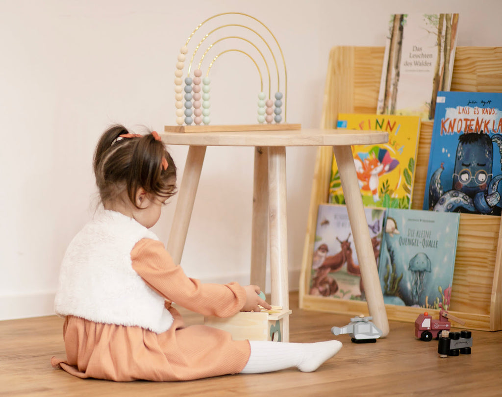 kleines Mädchen sitzt in der Bücherecke und spielt mit einem Steckspiel aus Holz mit Bücherregal und Kinderbüchern im Hintergrund