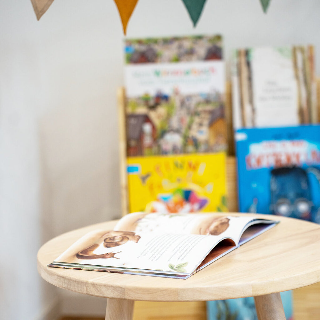 offenes Kinderbuch auf rundem Holztisch mit Bücherregal und Kinderbüchern im Hintergrund