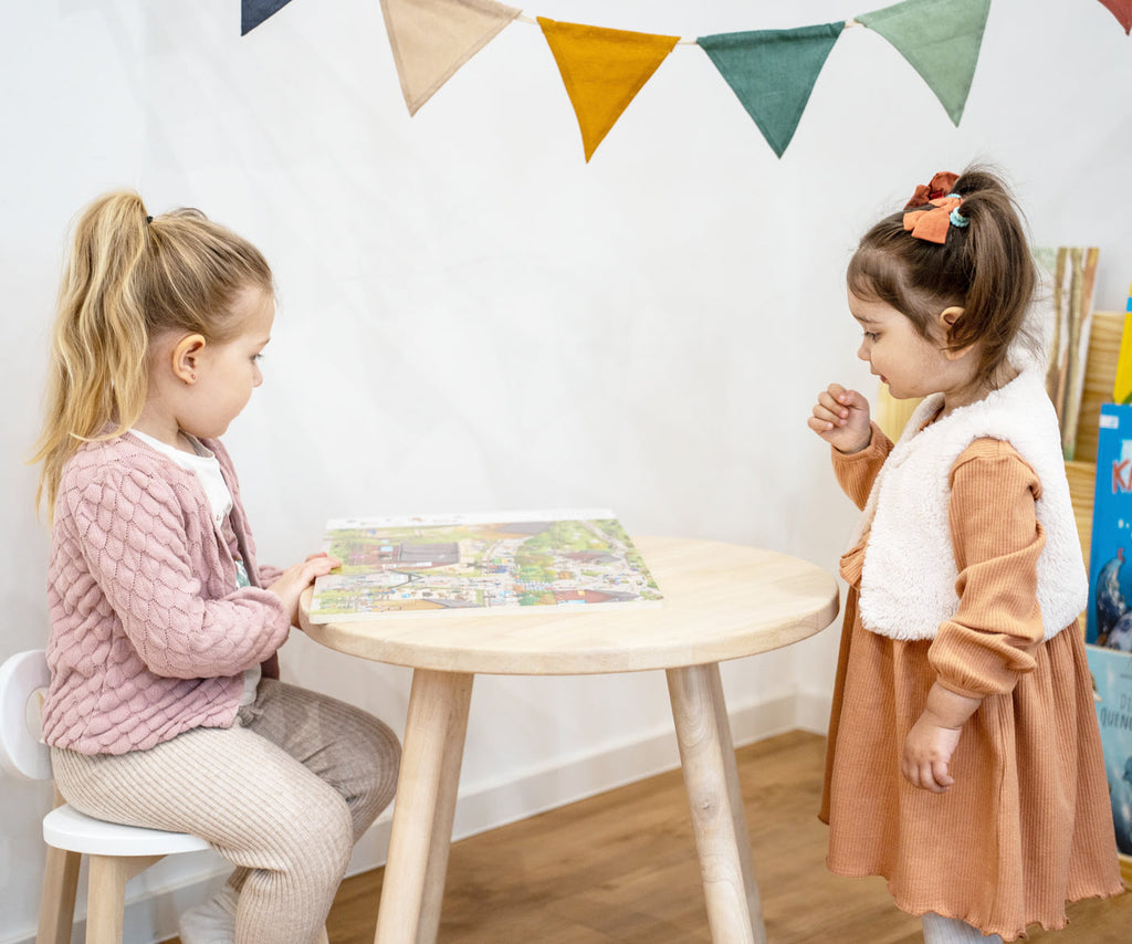 zwei Mädchen an einem runden Tisch aus Holz und schauen sich ein Wimmelbuch an