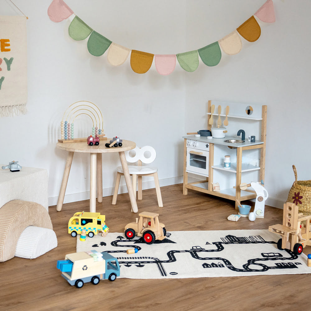 Kinderecke mit Kinderküche, Kindertisch und Stuhl aus Holz, Fahrzeuge aus Holz auf Spielteppich und Girlande in Pastelfarben
