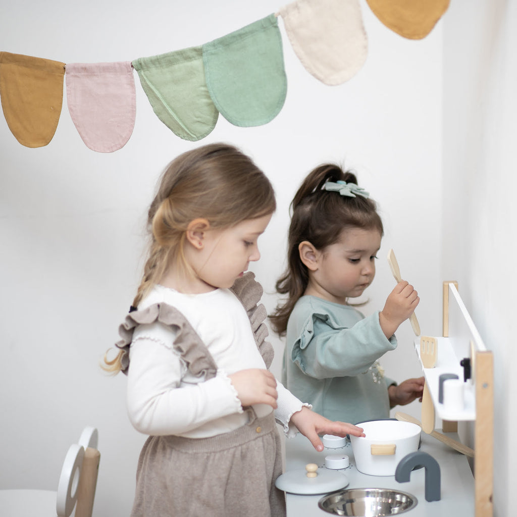 zwei Mädchen spielen in Kinderküche aus Holz in weiß unter Girlande in Pasteltönen