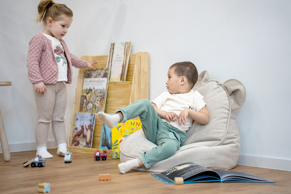 kleiner Junge sitzt im Sitzsack mit offenem Buch vor sich und Mädchen will ein Kinderbuch auf dem Bücherregal aus Holz nehmen