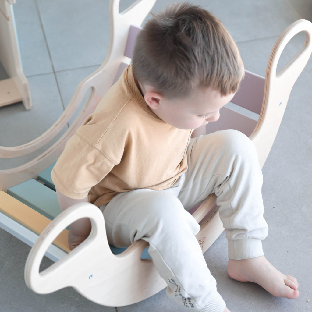 Kind sitzt im Kletterbogen Balance Wippe Holz Pastelfarben